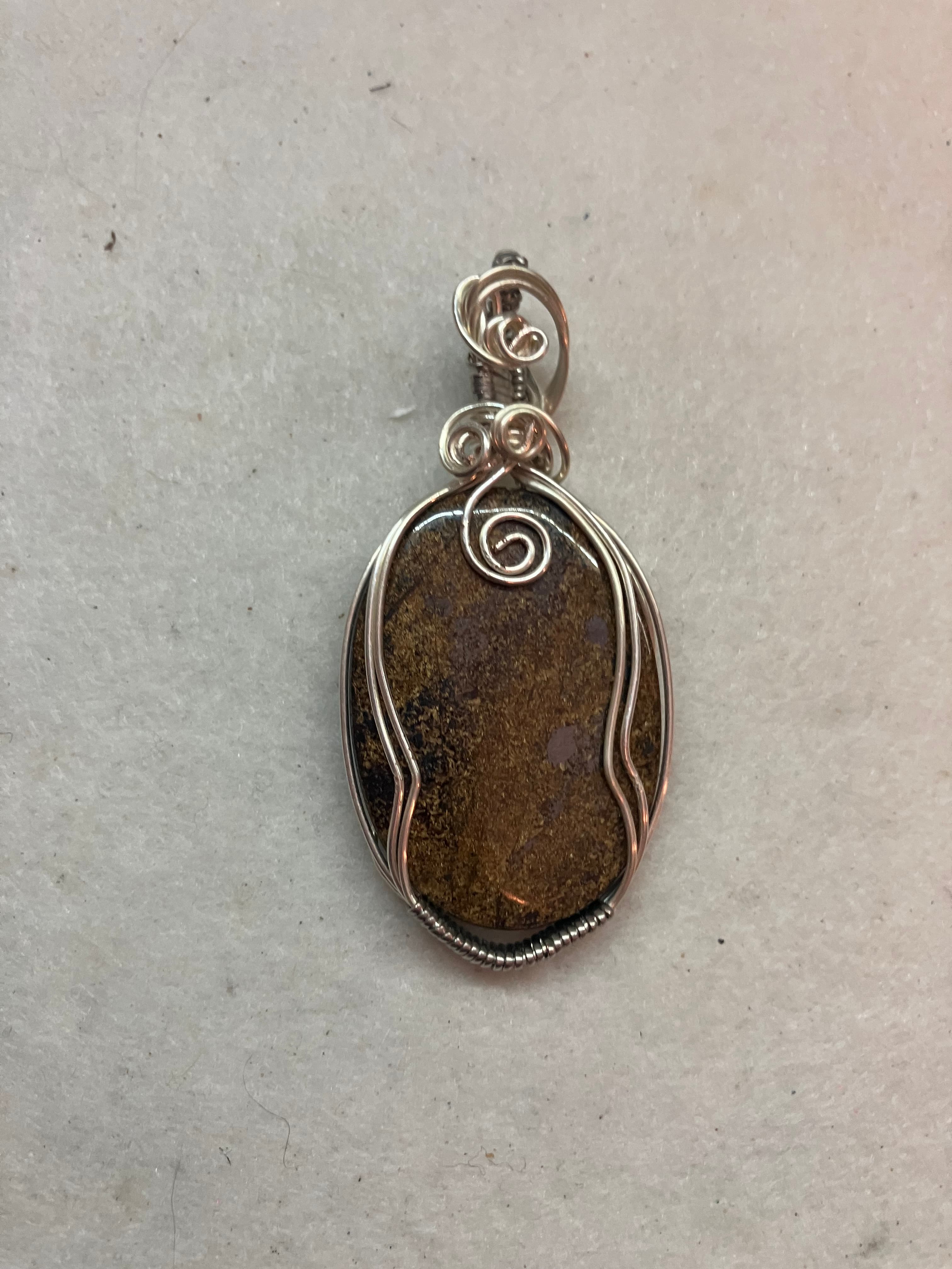 Oval brown stone pendant intricately wrapped in silver wire with decorative swirls and loops.