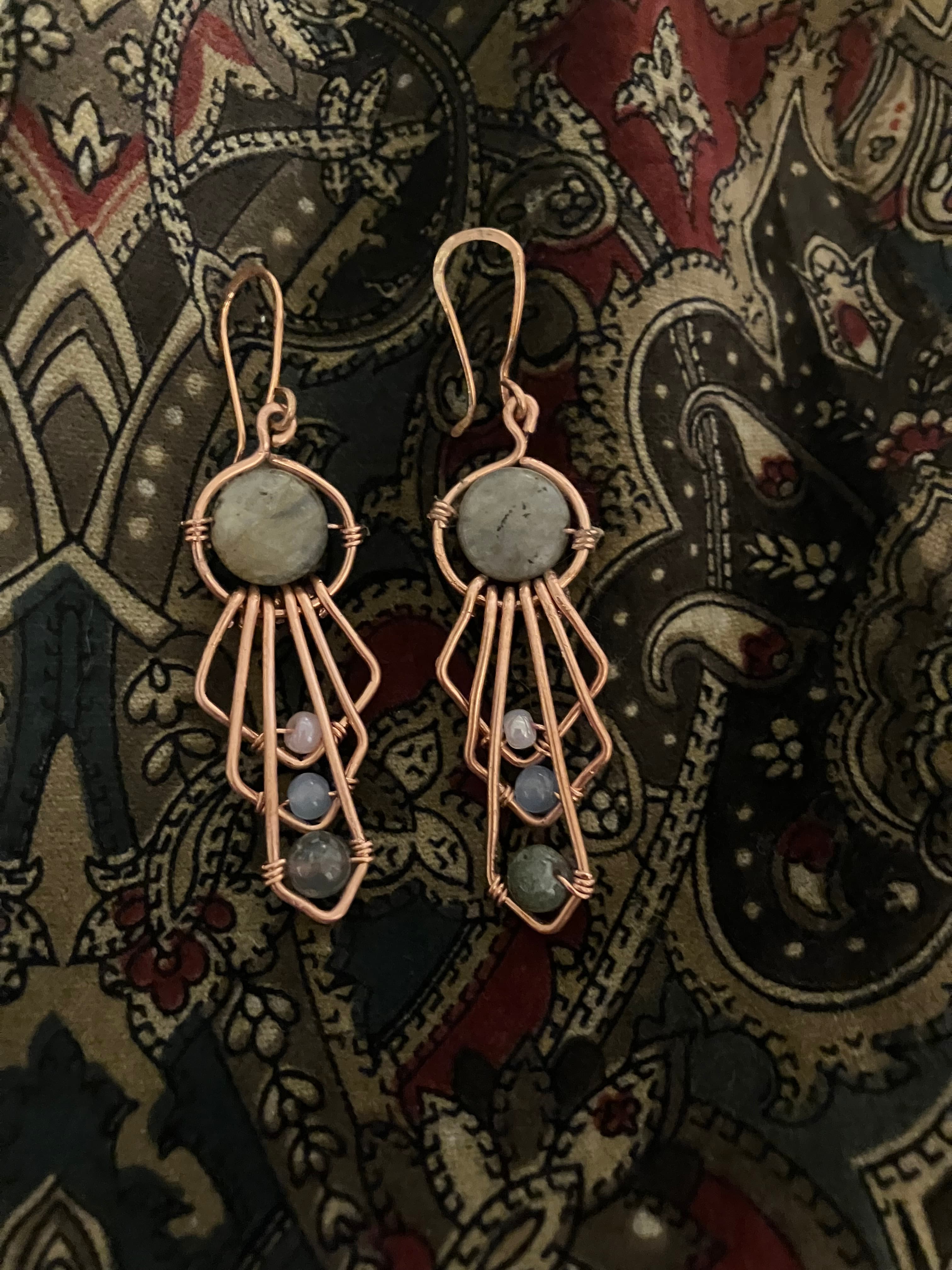 Handmade copper wire earrings with grey stones and beads resting on a paisley patterned fabric.