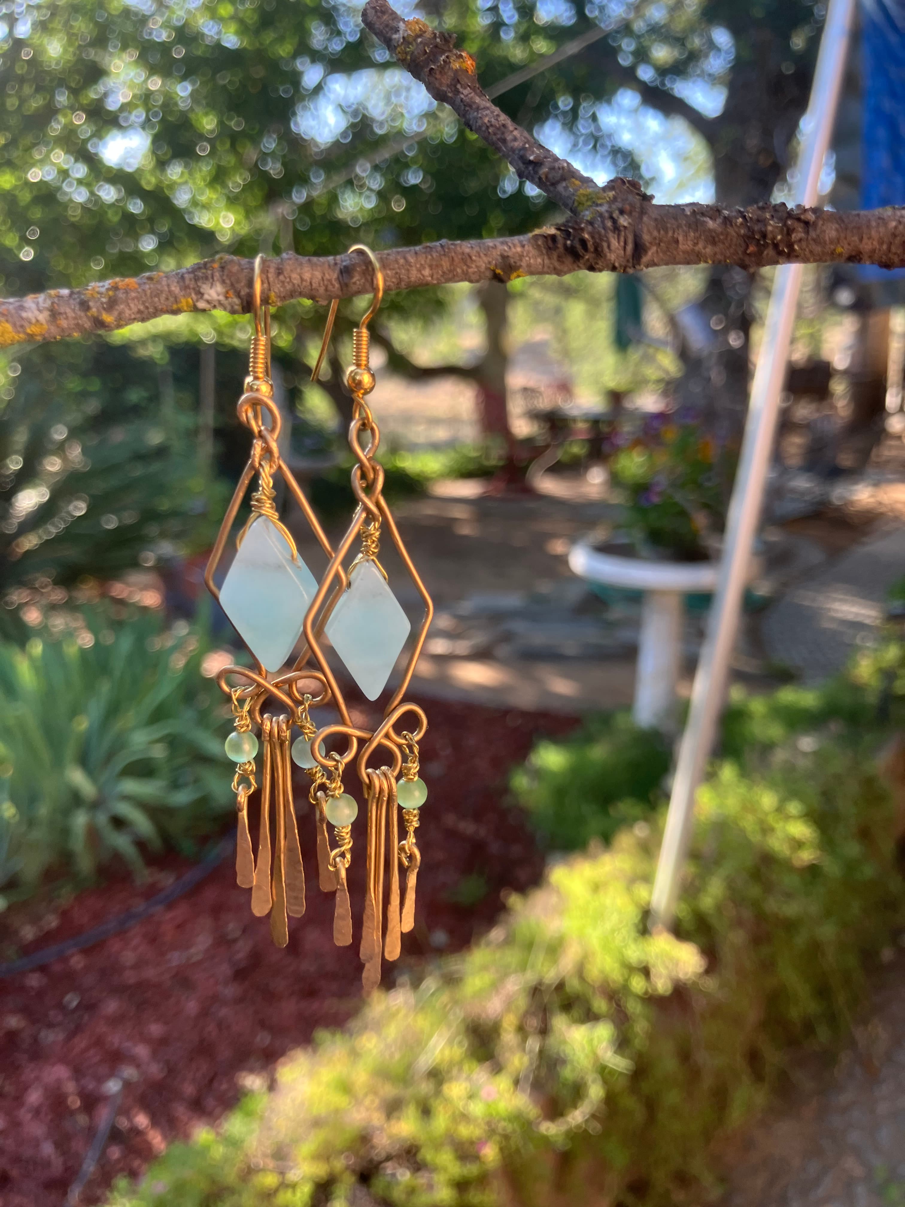 Gold chandelier earrings with light blue diamond stones and hammered dangles hanging from a branch.