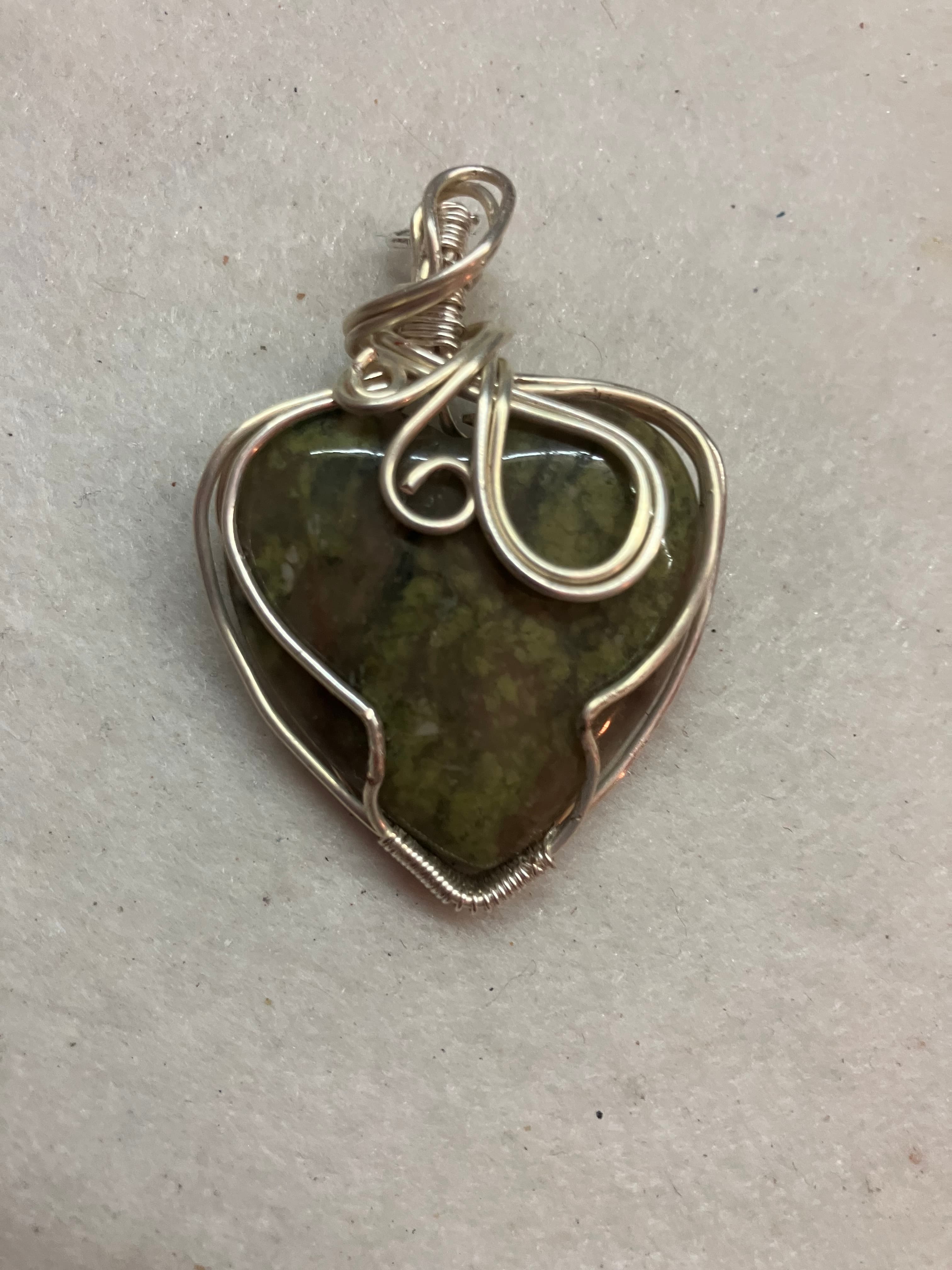 Heart-shaped green and brown stone pendant intricately wrapped in decorative silver wire on grey background.